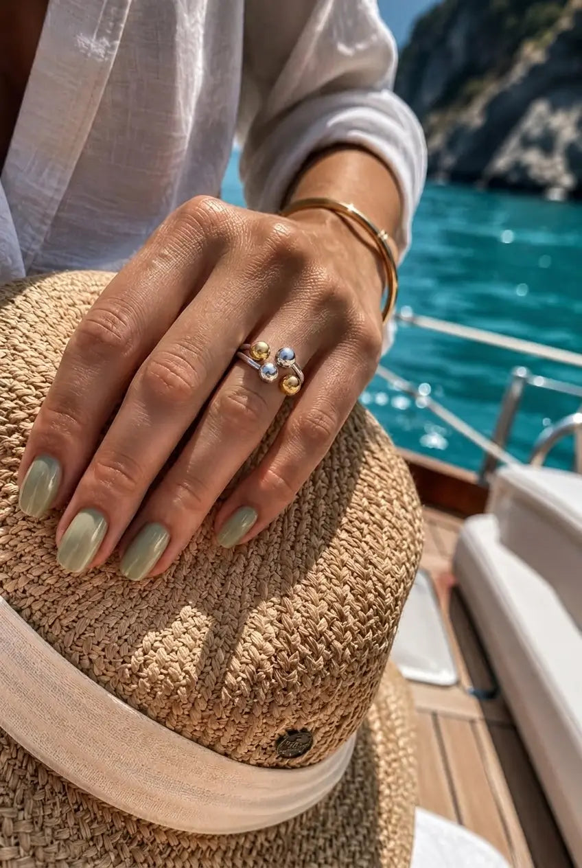 A close-up lifestyle shot of a woman's hand resting on a straw hat, wearing a gold ring with a marquise-cut gemstone and round cubic zirconia stones. The background shows a sunny day on a boat with turquoise water and rocky cliffs.
