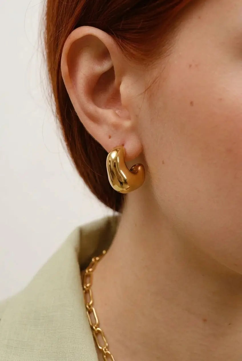 A close-up of a Model's ear wearing a chunky, polished gold crescent-shaped hoop earring, with a gold chain necklace and a light sage green top visible.