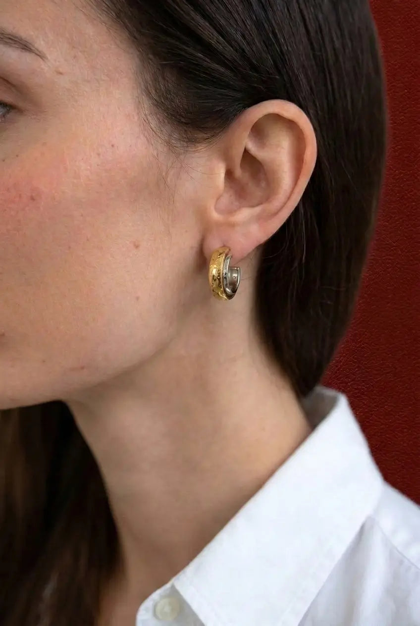 A close-up side profile of a woman wearing a small gold hoop earring with a textured, hammered finish, paired with a white collared shirt against a dark red background.