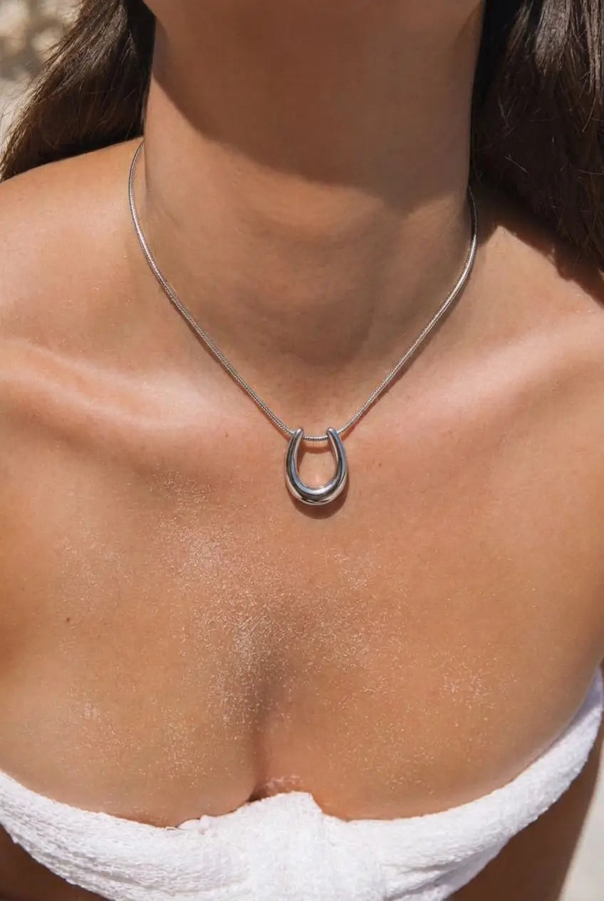 A close-up of a model wearing a silver horseshoe pendant necklace on a delicate chain, styled with a white strapless top.