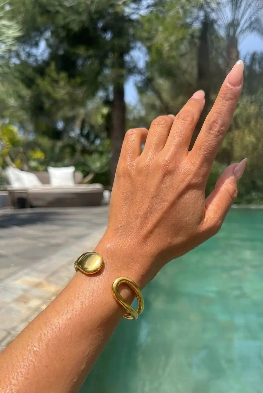 A gold-toned double-band cuff bracelet worn on a woman's wrist, shown in a sunny outdoor setting with a swimming pool and green trees in the background.