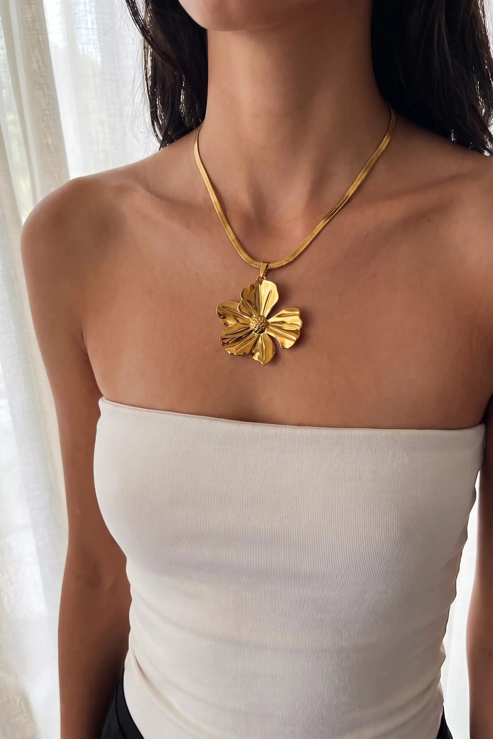 A close-up of a model wearing a textured gold flower pendant necklace with a matching gold chain, paired with a white strapless top.