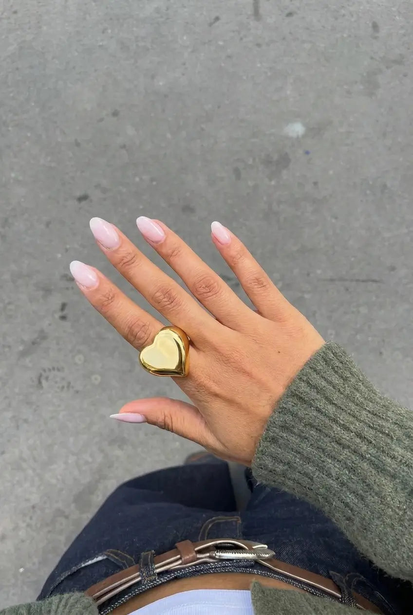 A gold heart-shaped ring worn on a person's hand, shown in a top-down view against a gray concrete pavement background.