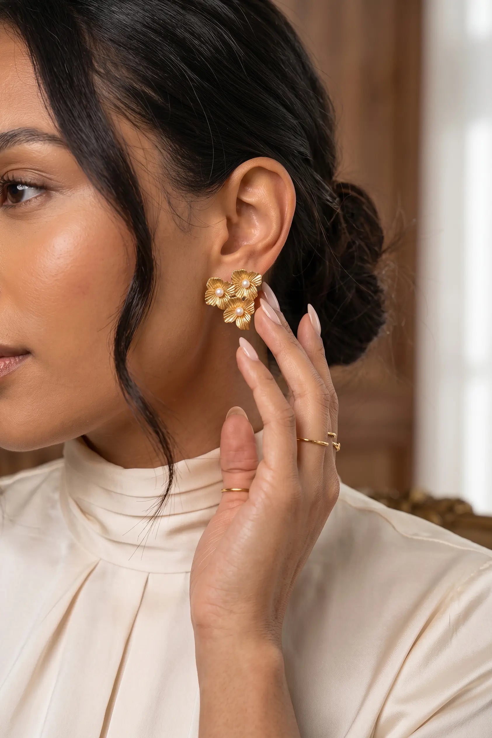 A close-up lifestyle shot of a woman wearing a gold floral cluster stud earring and a gold ring, wearing a cream-colored high-neck blouse.