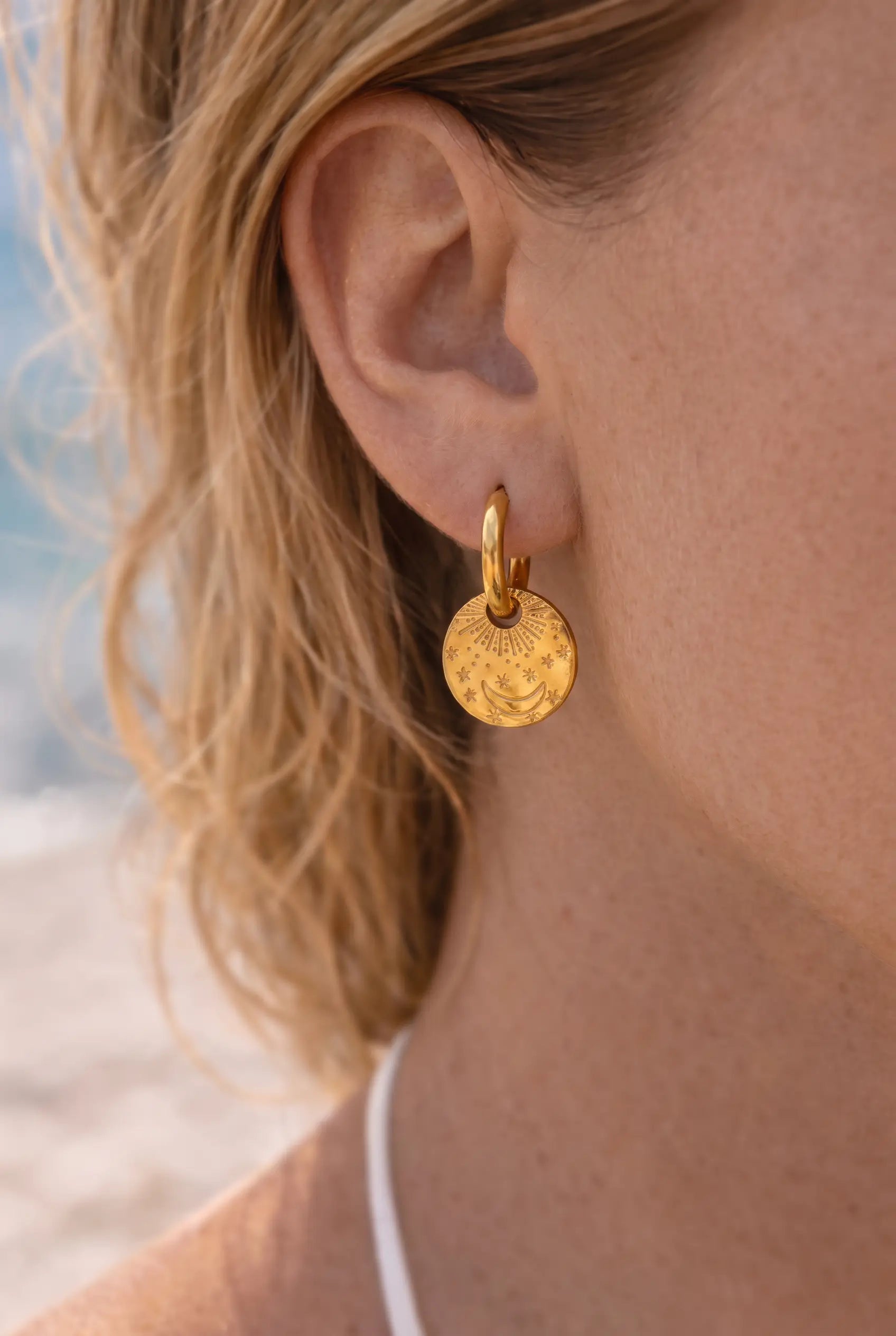 A close-up of a woman's ear wearing a small gold hoop earring with a textured, hammered circular disc pendant.