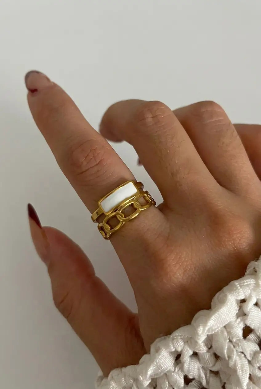 A close-up of a hand wearing a gold chain-link ring with a white inlay, paired with a white knit sleeve against a plain light background.