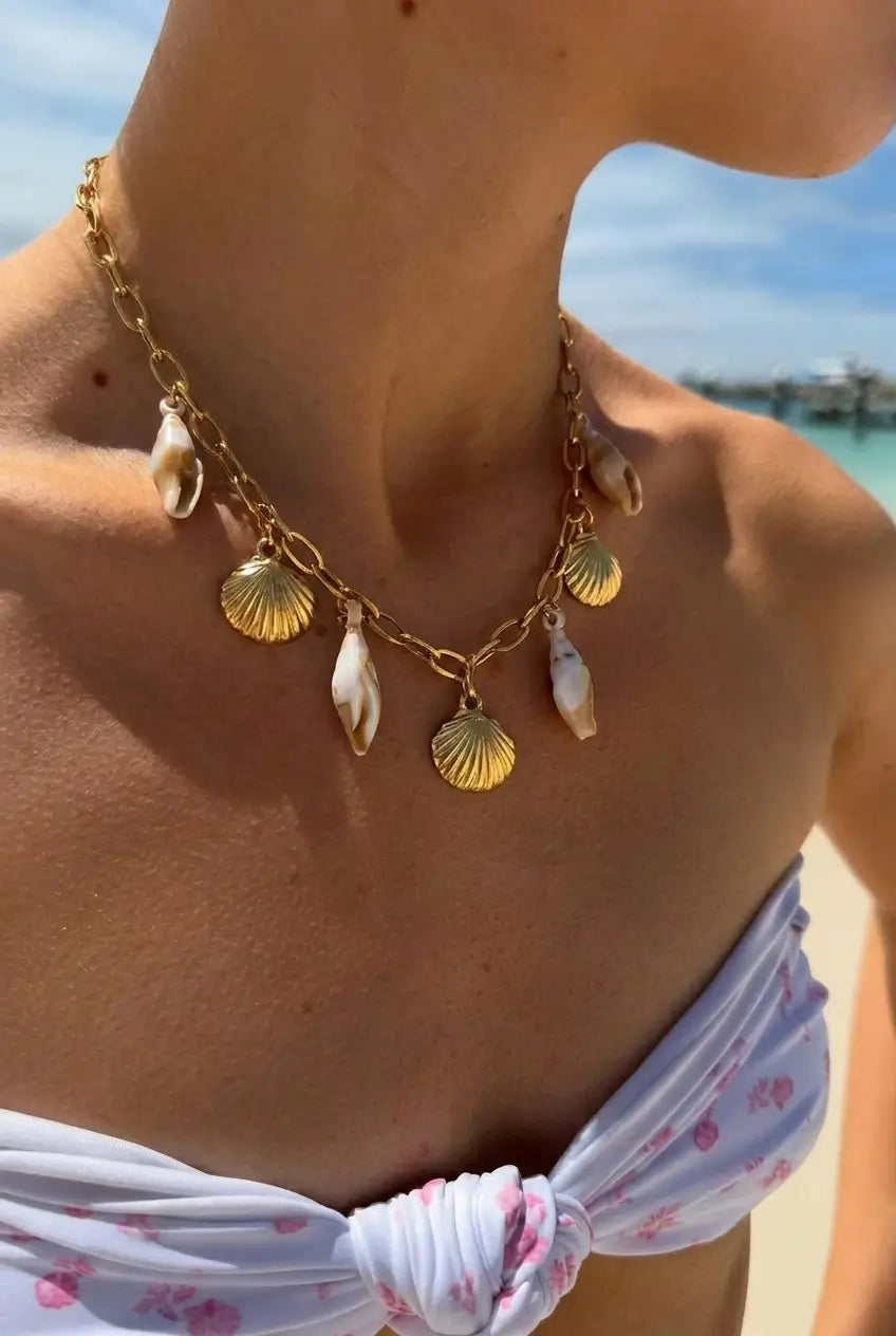 A close-up lifestyle shot of a woman wearing a gold chain necklace with dangling seashell charms, set against a sunny beach background.
