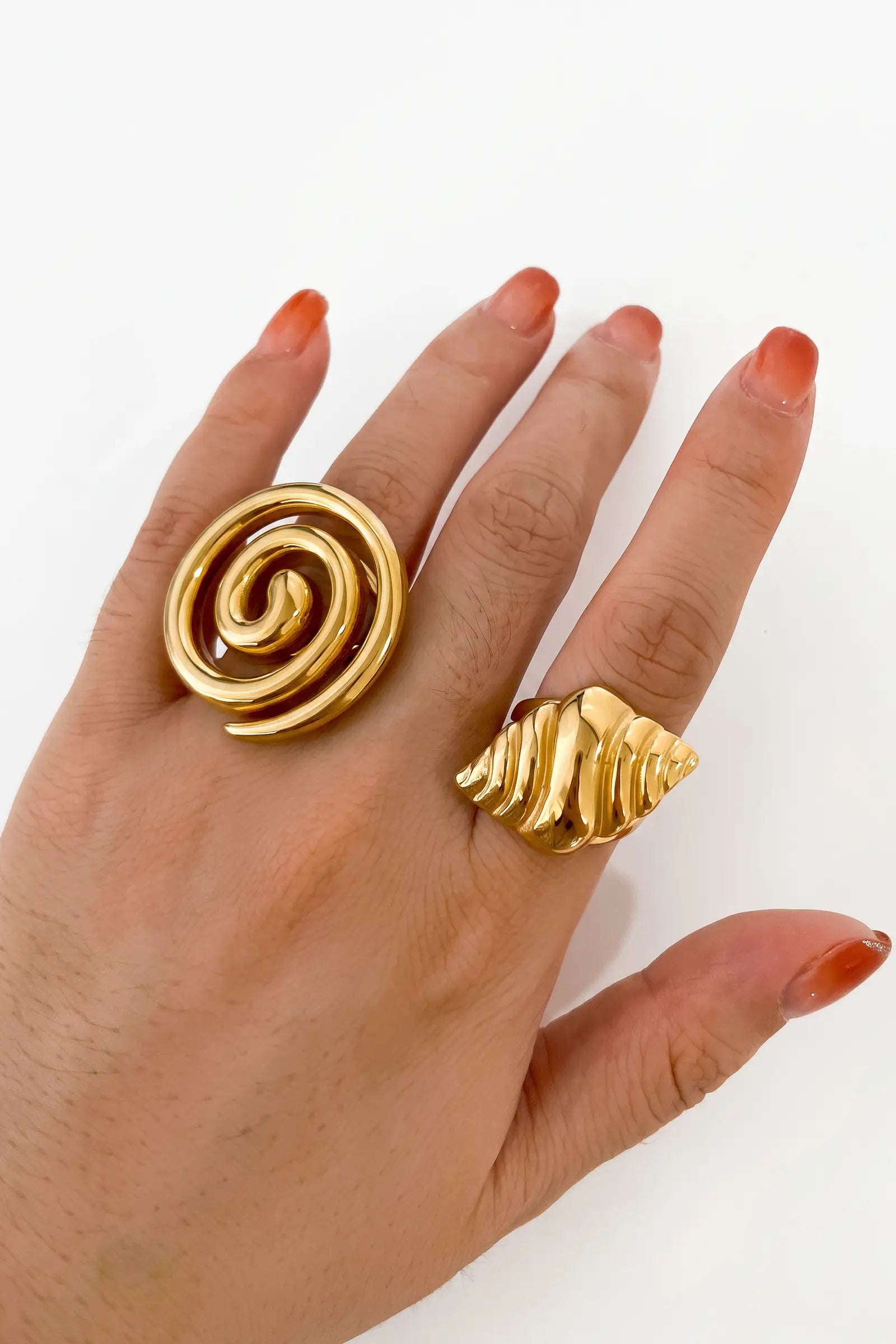 A close-up of a hand wearing two chunky gold rings, one with a spiral design and the other with a textured shell-like layered design, against a plain white background.