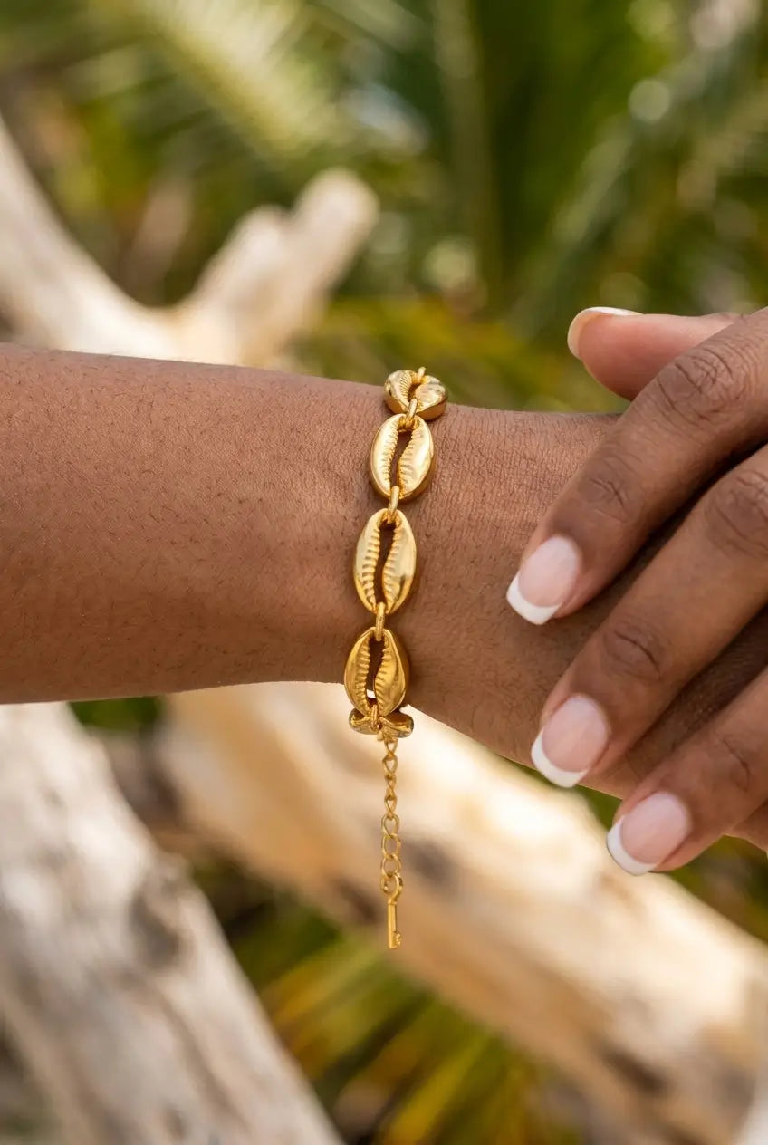 A gold chain bracelet with textured shell links worn on a person's wrist, shown against a blurred tropical background with palm leaves and driftwood.