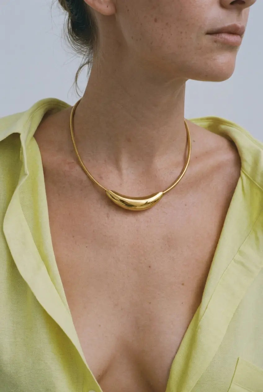 A close-up of a model wearing a polished gold crescent-shaped necklace with a wide collar of a pale yellow-green shirt.