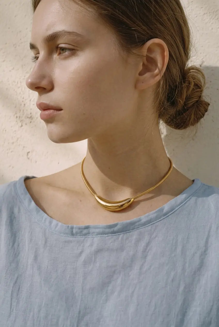 A woman in profile wearing a light blue linen top and a gold-toned layered choker necklace, set against a textured beige wall.