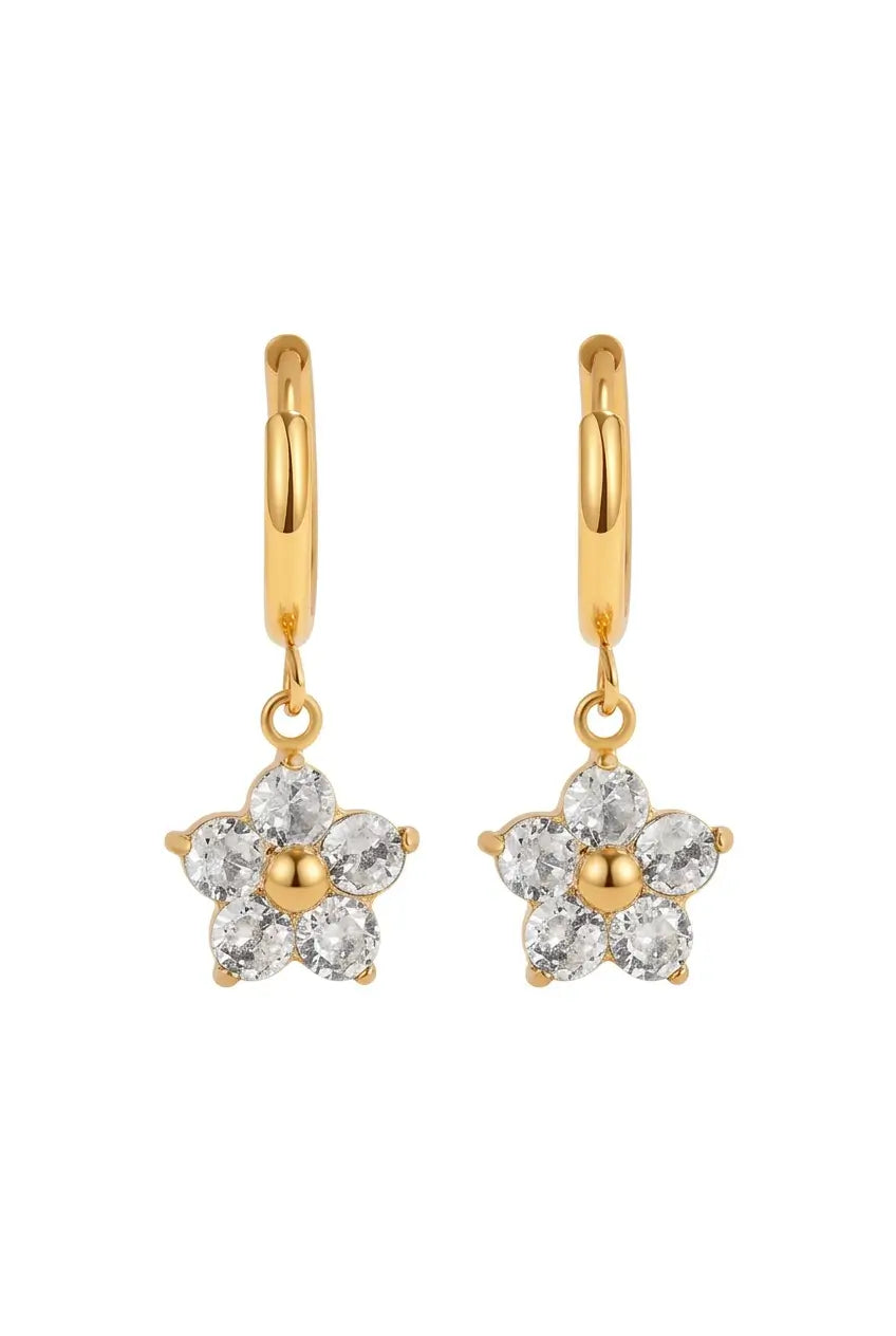 A pair of gold huggie hoop earrings with dangling flower charms. Each flower charm features six clear round-cut stones surrounding a small gold center, set against a plain white background.