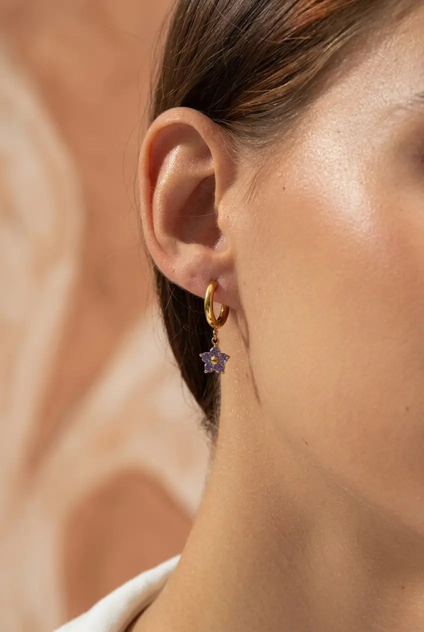 A close-up of a model's ear wearing a small gold hoop earring with a dangling purple star-shaped charm.