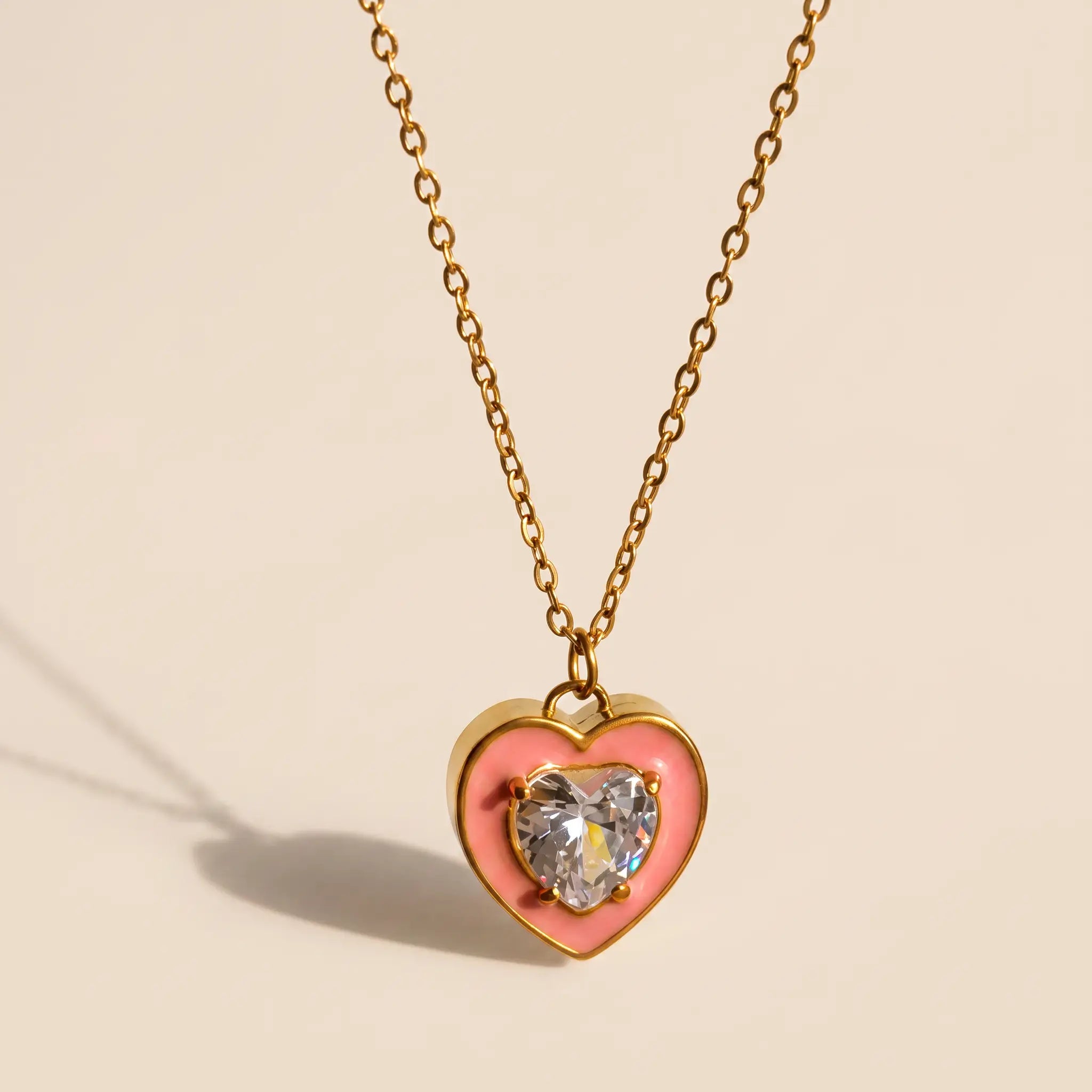 A gold heart pendant necklace featuring a pink enamel heart and a clear heart-shaped gemstone in the center, suspended from a delicate gold chain against a warm beige background.