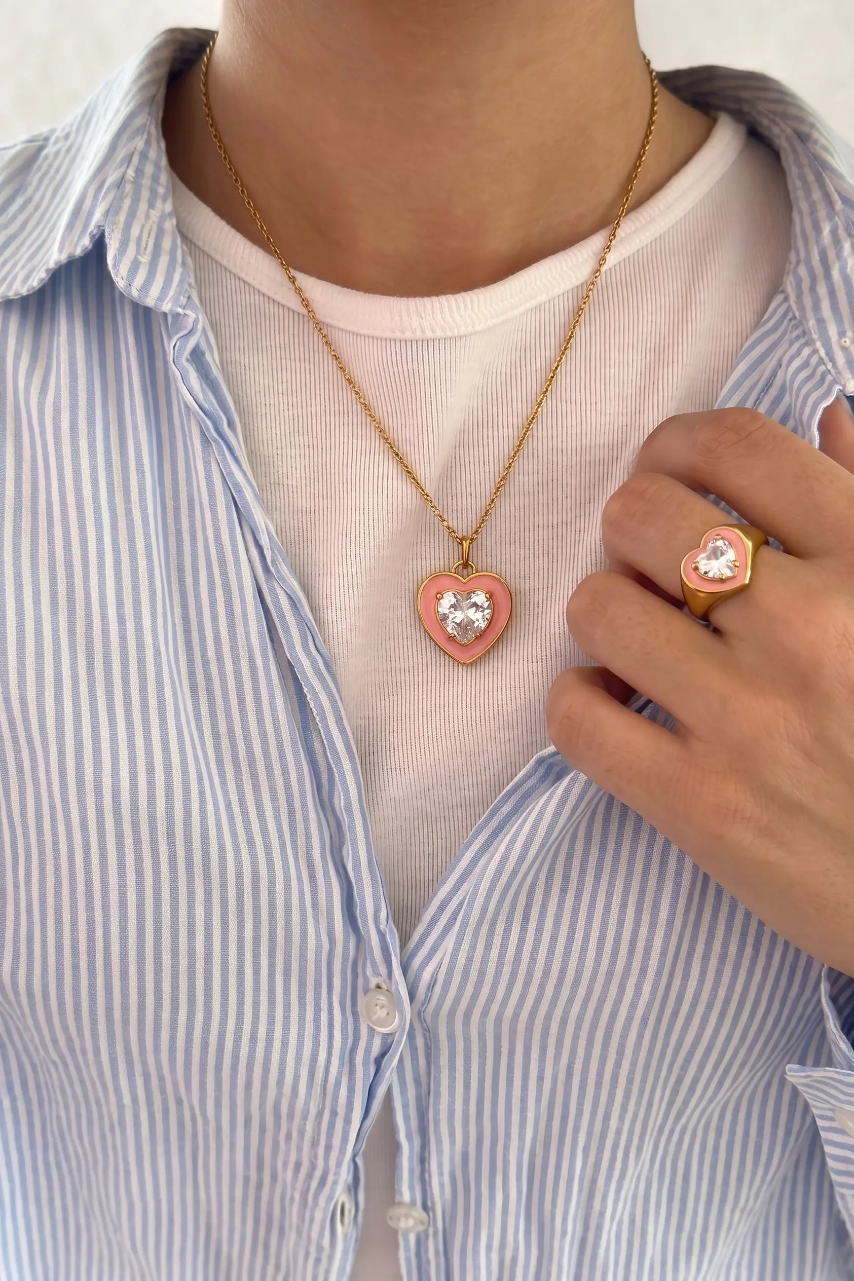 A close-up of a person wearing a gold heart pendant necklace and a matching heart ring, styled with a white t-shirt and a blue and white striped button-down shirt.