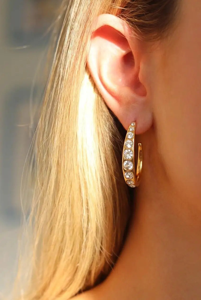 A close-up of a model's ear wearing a gold crescent-shaped hoop earring adorned with clear pavé stones, with warm sunlight highlighting the hair and jewelry.