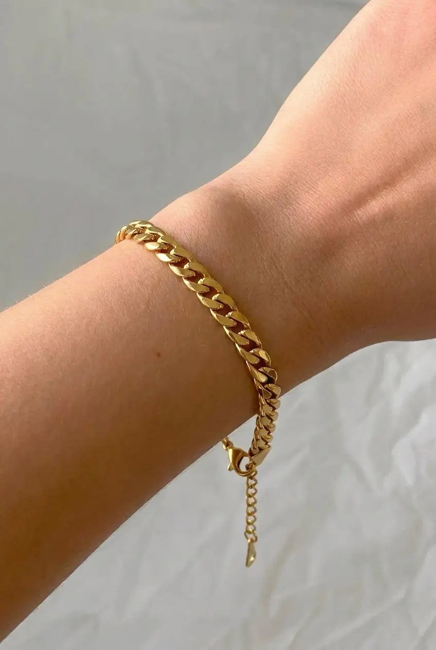 A gold-toned chunky curb chain bracelet worn on a person's wrist, shown against a light gray fabric background.