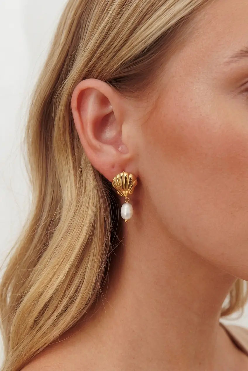 A close-up of a woman's ear wearing a gold scallop-shaped stud earring with a dangling white pearl.