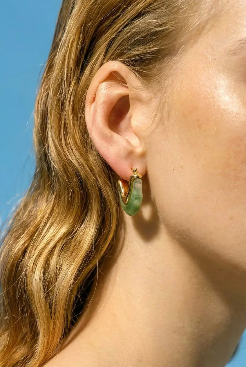 A close-up of a model's ear wearing a small gold hoop earring with a green jadeite crescent drop pendant, set against a solid blue background.