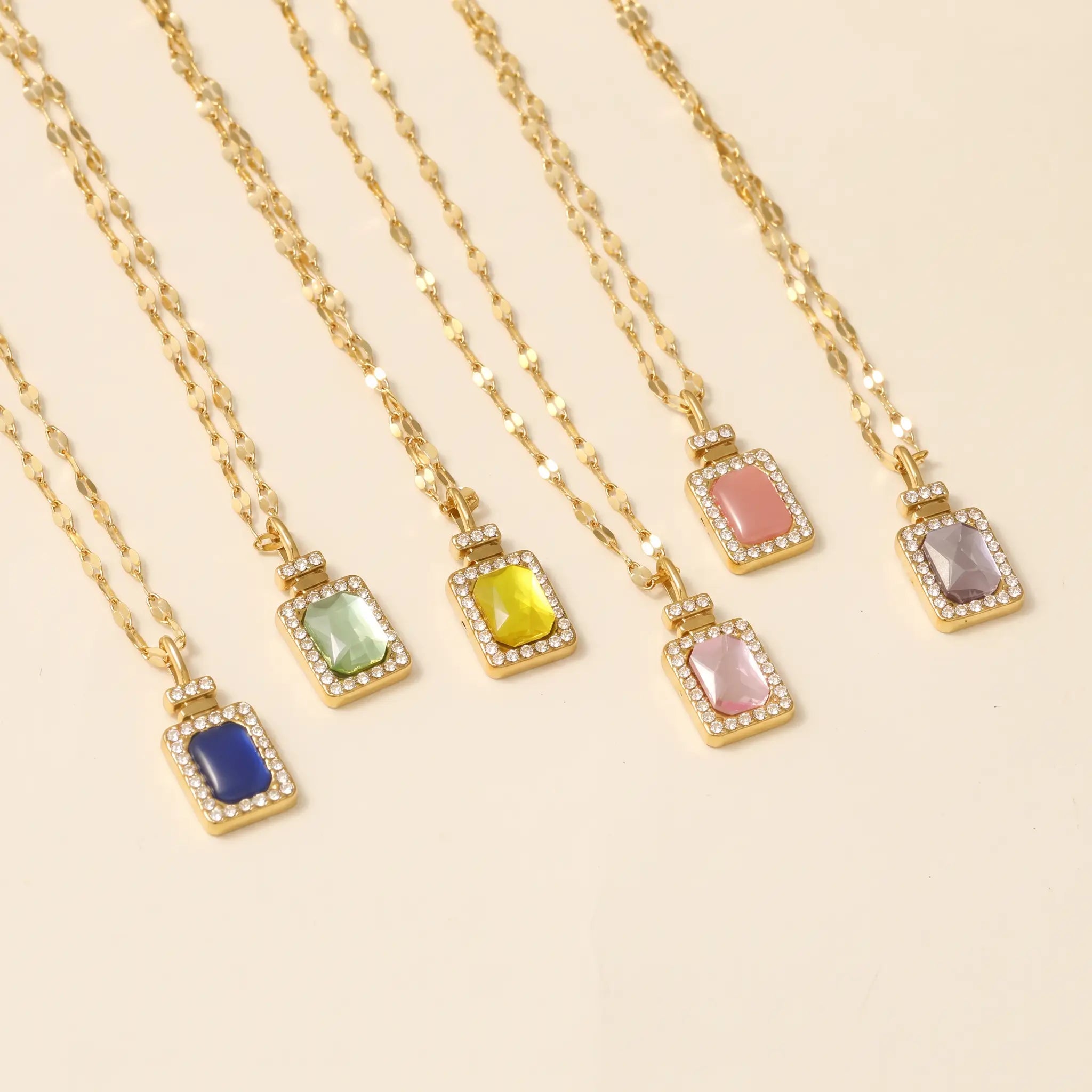 Five gold chain necklaces with rectangular pendant frames, each set with a different colored stone (blue, green, yellow, pink, and lavender) and small clear rhinestones, arranged diagonally on a light beige background.