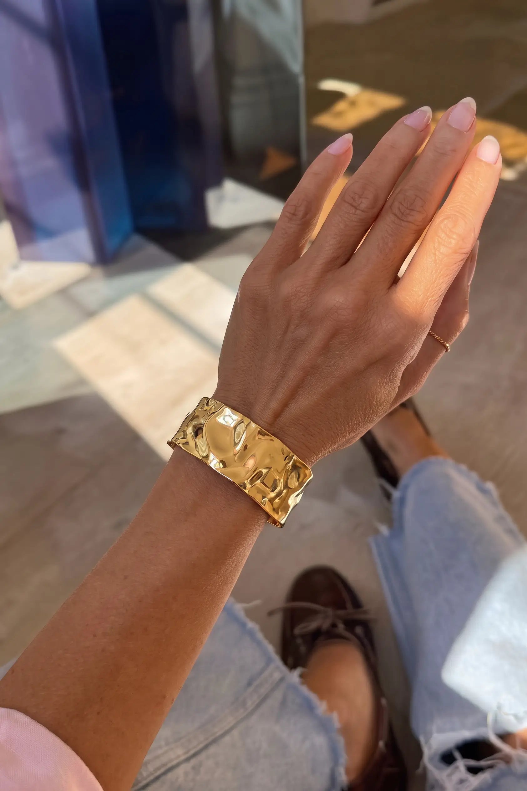 A person wearing a chunky, textured gold cuff bracelet on their wrist, shown in a close-up lifestyle shot with denim jeans and brown shoes.