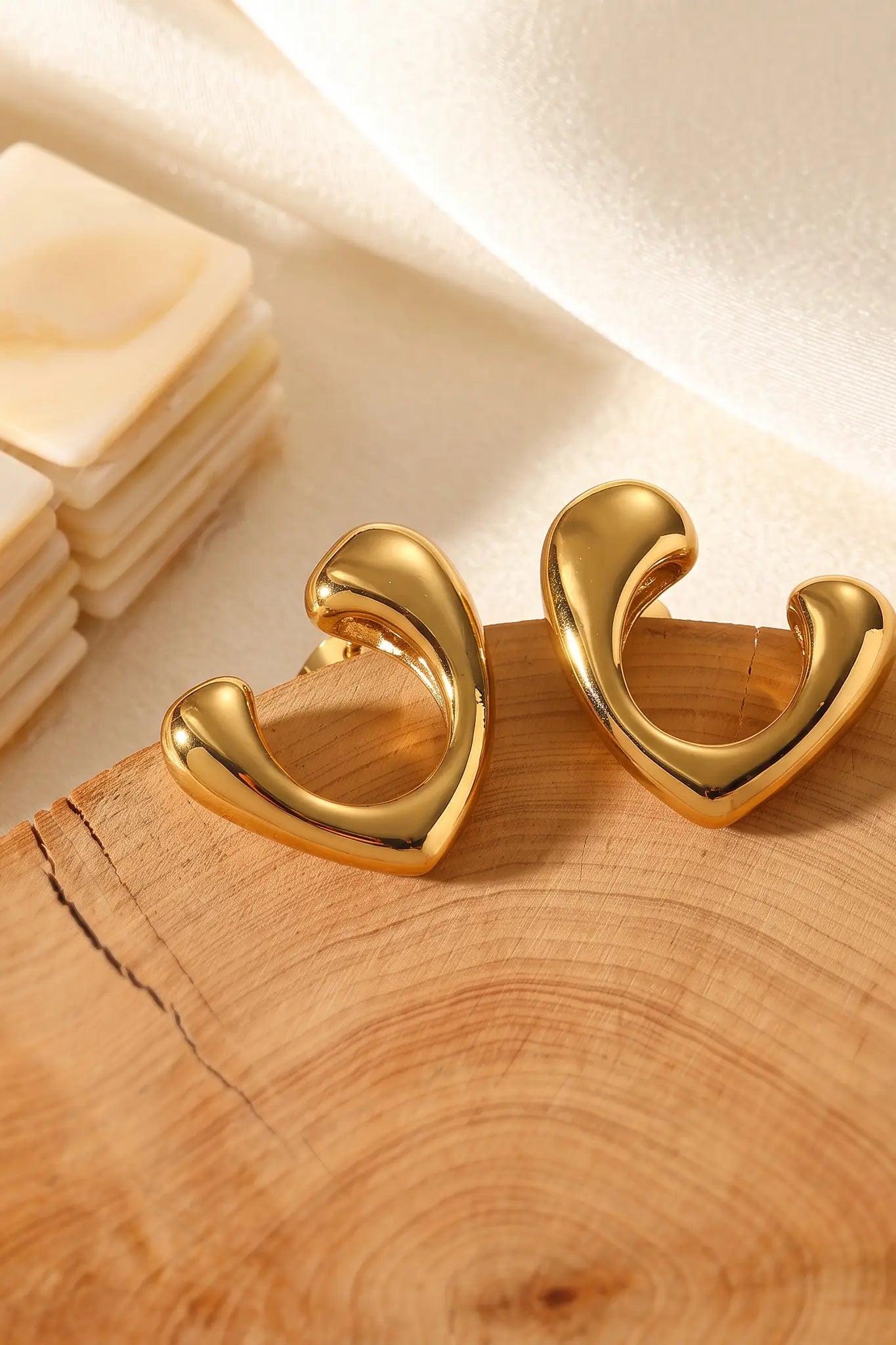 A pair of gold-toned, open heart-shaped hoop earrings displayed on a rustic wooden slice with a soft white fabric background.