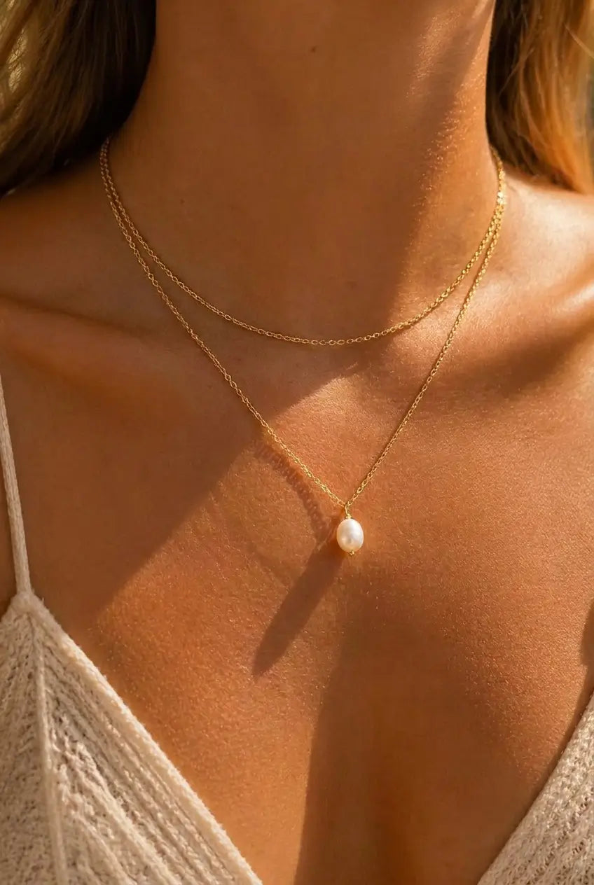 A close-up of a model wearing a delicate double-layered gold necklace with a single white pearl pendant, styled with a white lace top.