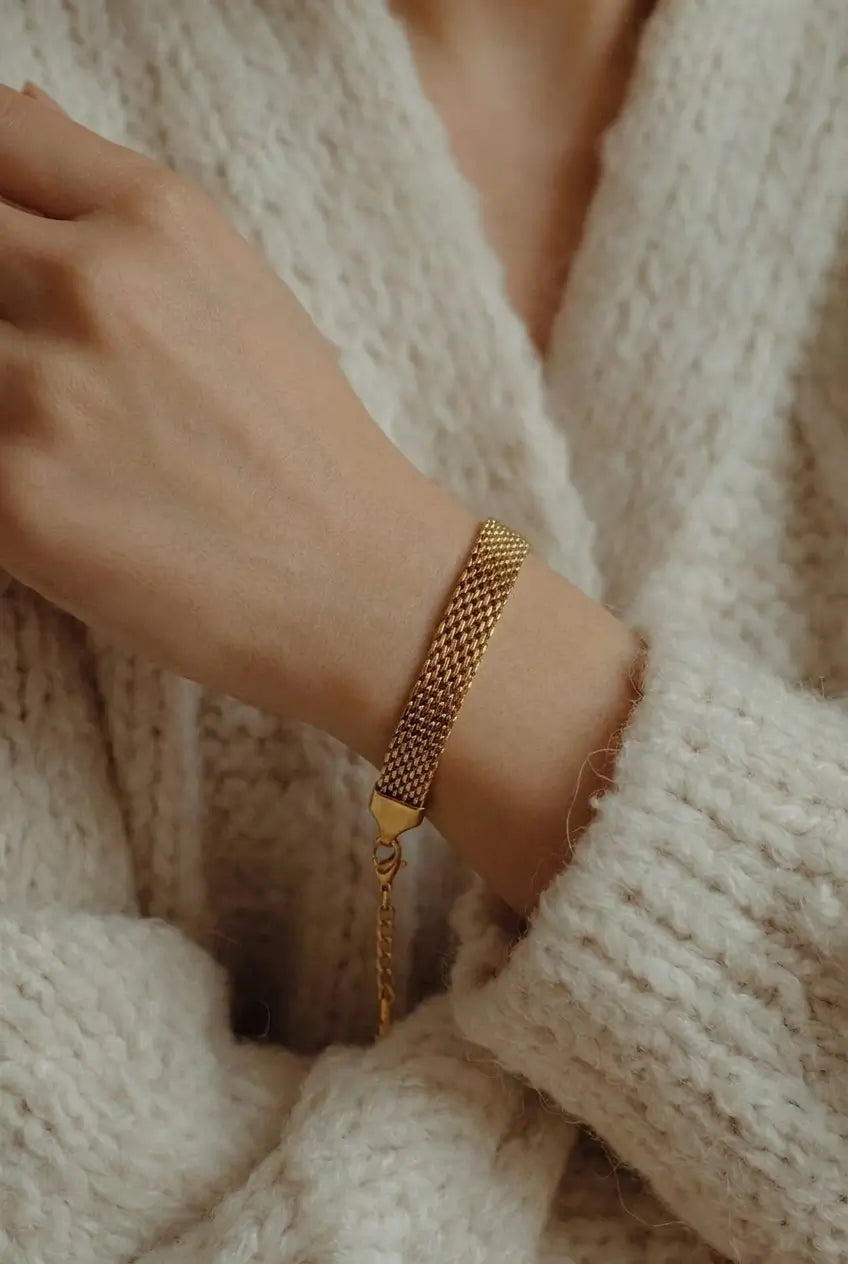 A close-up of a person's wrist wearing a gold mesh bracelet, styled with a chunky cream-colored knit cardigan.