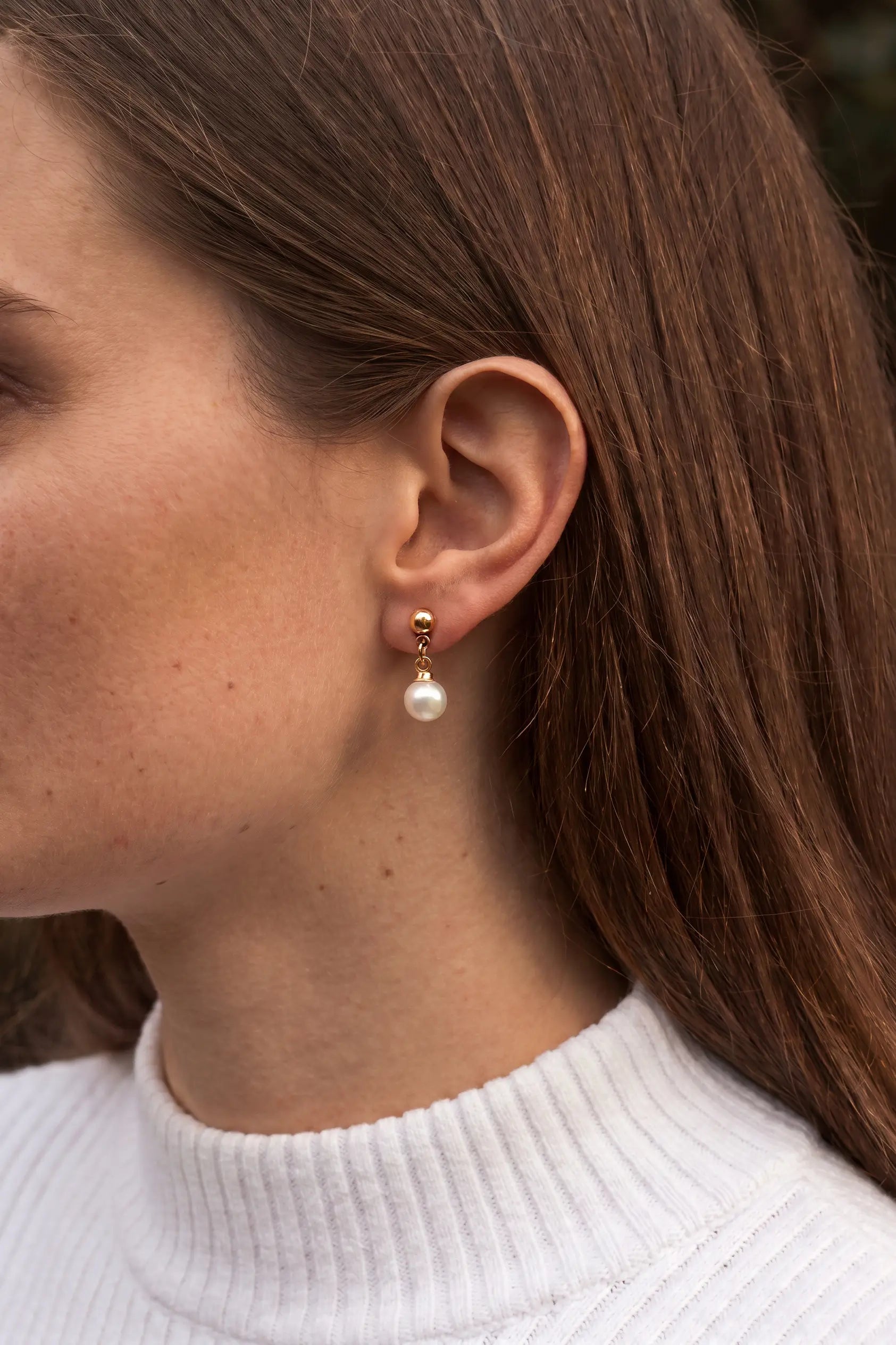 A close-up side profile of a woman wearing a small gold hoop earring with a dangling white pearl, paired with a white ribbed turtleneck sweater.