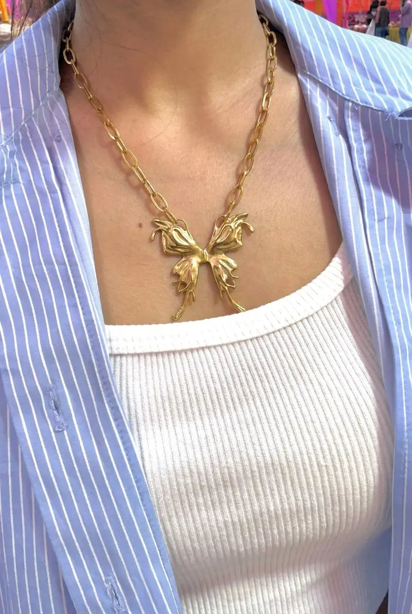 A gold butterfly pendant necklace worn by a person, styled over a white ribbed tank top and a light blue and white striped button-down shirt.