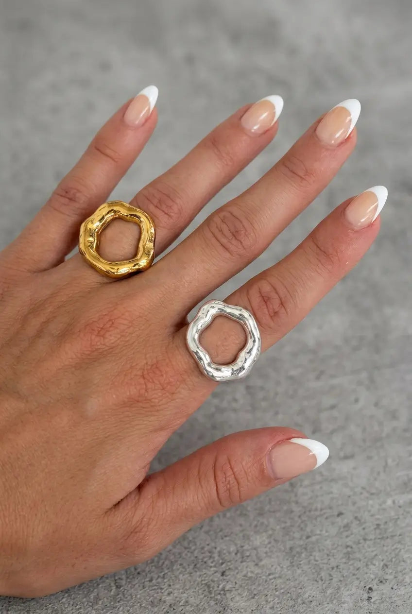 A hand with a French manicure wearing two textured, ring-shaped open pendants, one in polished gold and the other in polished silver, set against a neutral gray background.
