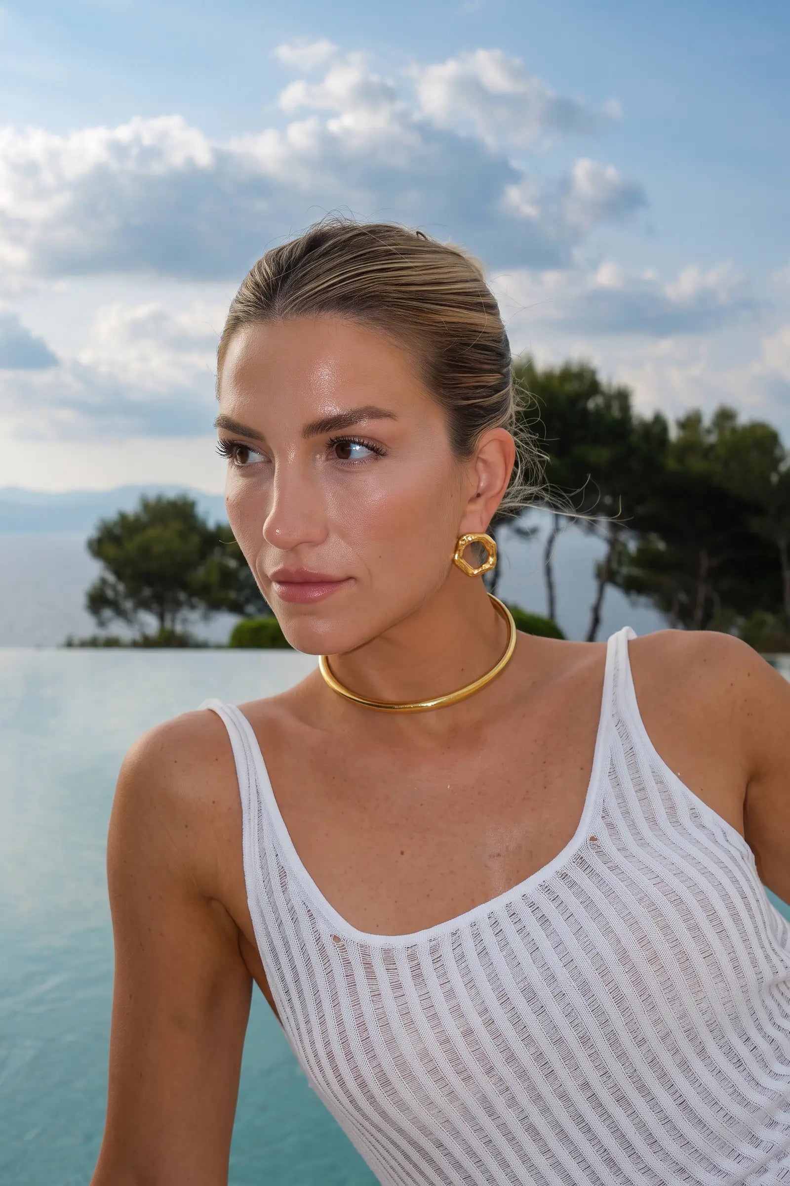 A woman with her hair pulled back wears a white ribbed tank top and a gold choker necklace, standing by an infinity pool with a blue sky and trees in the background.
