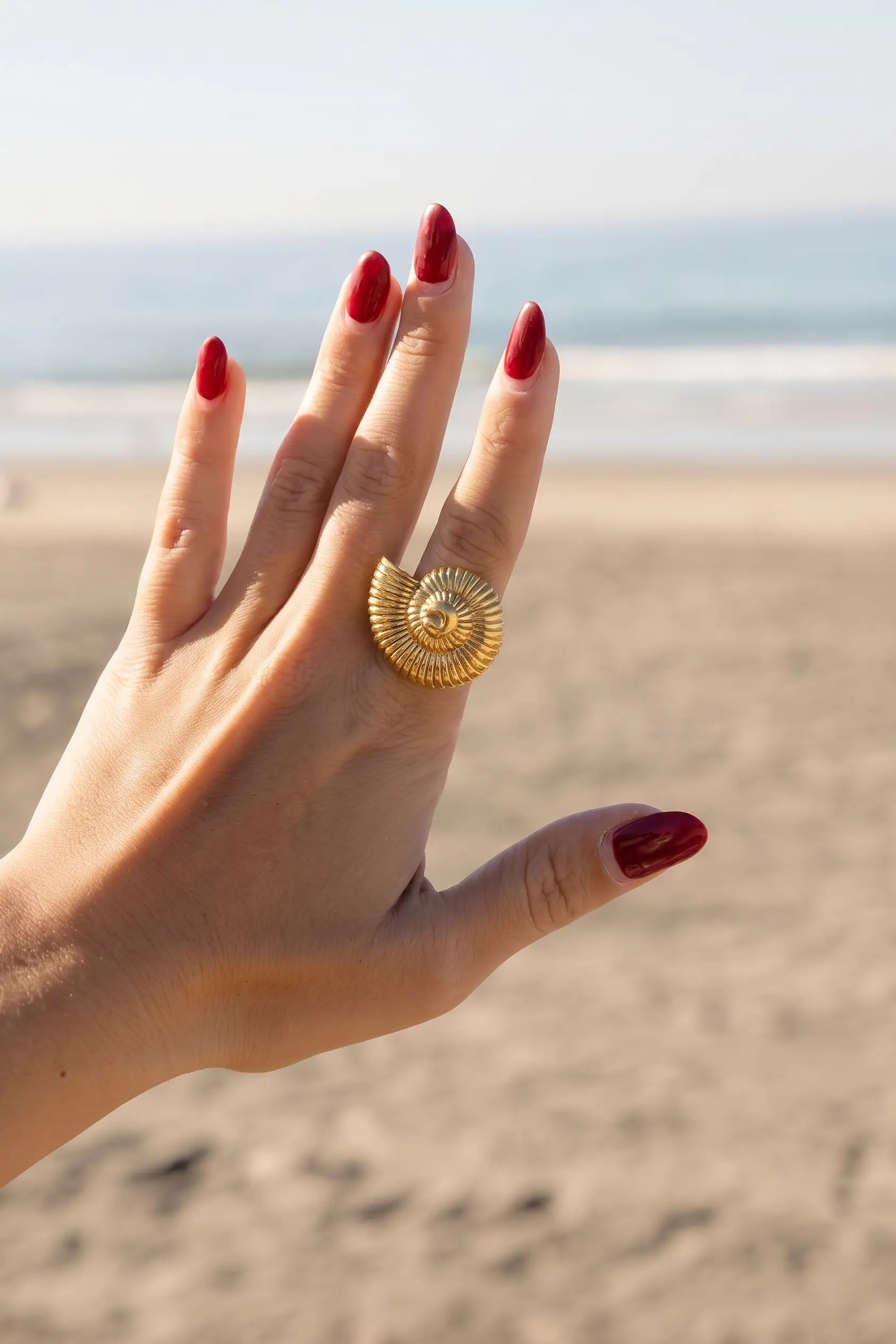 A gold spiral shell ring worn on a hand with red manicured nails, set against a blurred beach background.