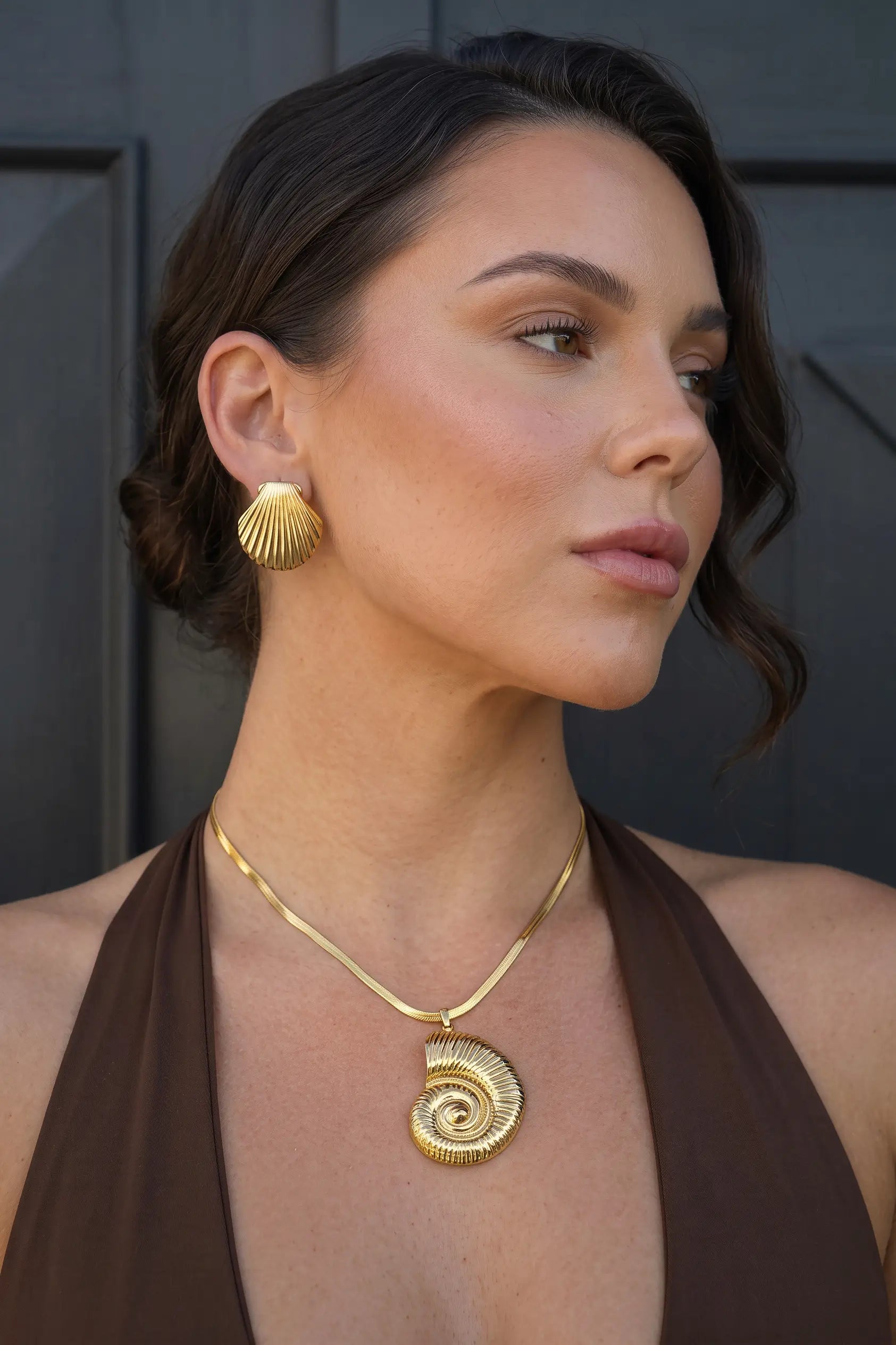 A woman wearing a gold spiral pendant necklace and matching gold shell-shaped drop earrings, shown in a close-up portrait against a dark gray background.