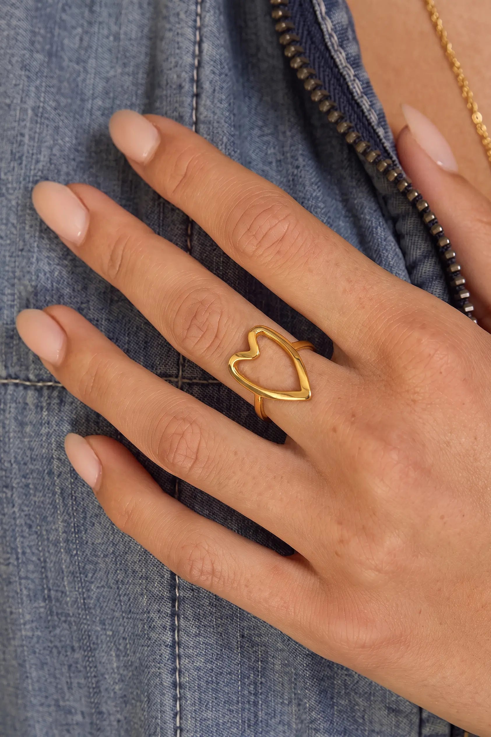 A close-up of a hand wearing a delicate gold ring with an open heart design, resting on a blue denim jacket.