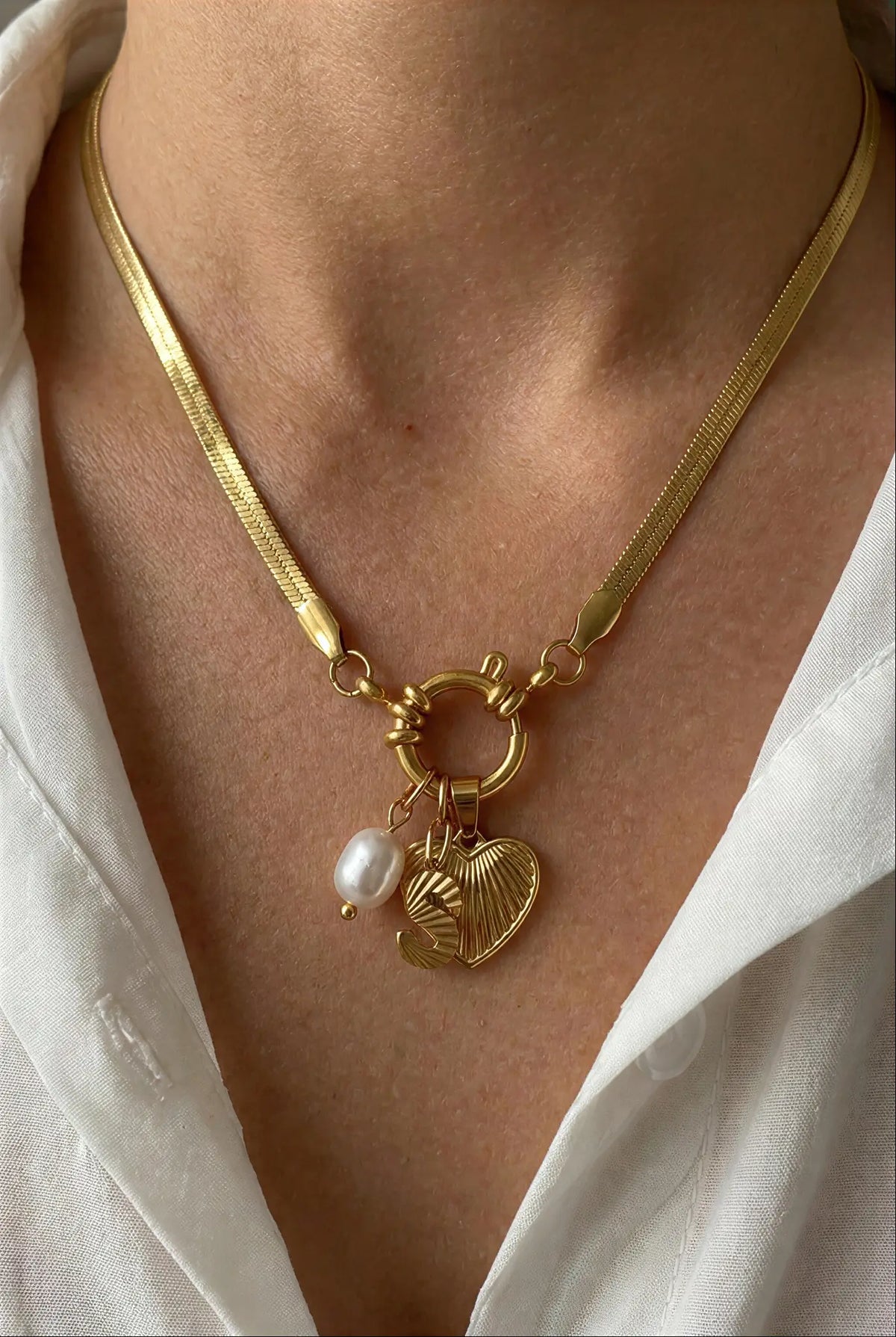 A close-up of a gold chain necklace with a toggle clasp, featuring a textured heart charm, a small leaf charm, and a white pearl bead, worn by a person in a white collared shirt.
