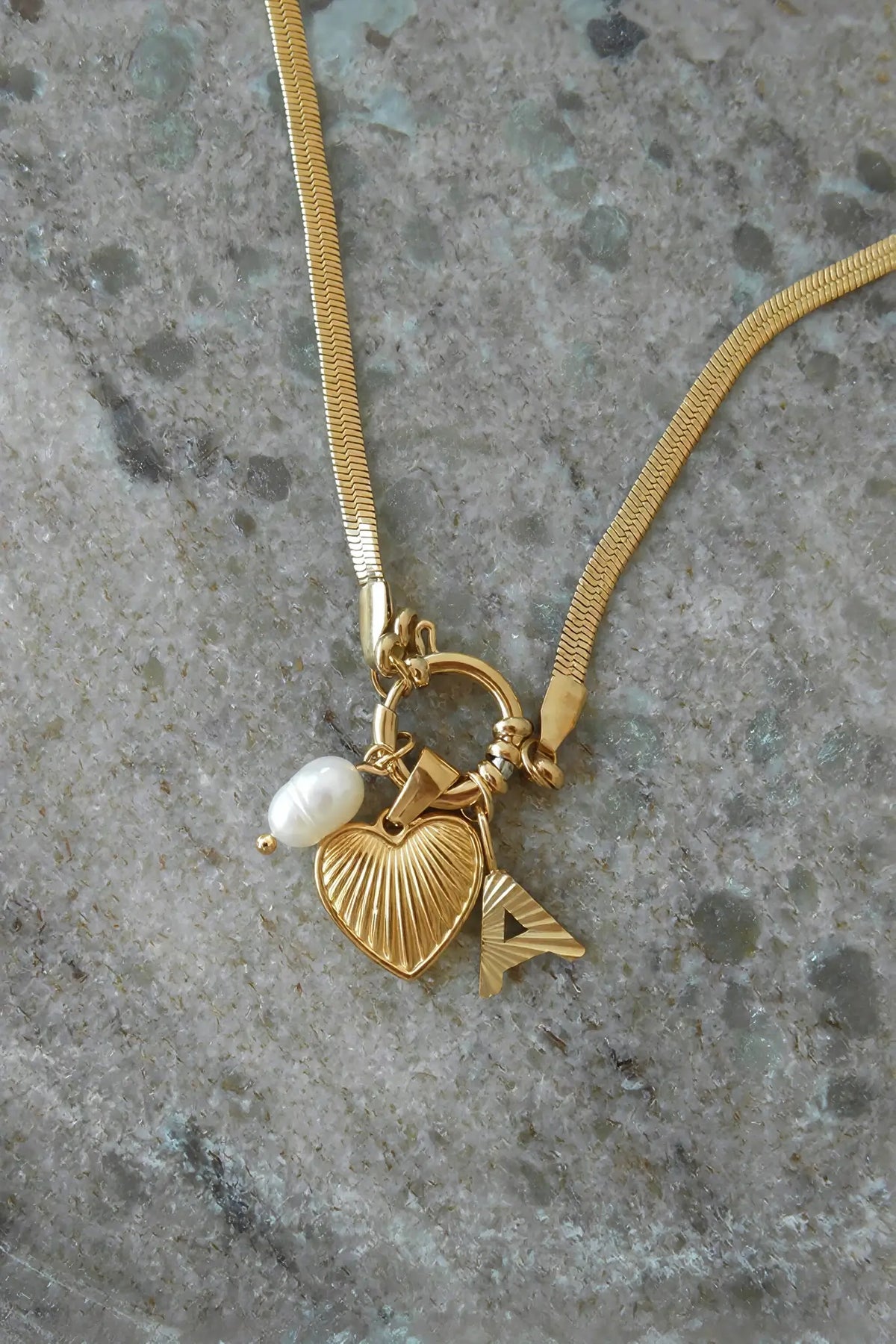A gold snake chain necklace with a heart-shaped pendant, a small white pearl bead, and a gold letter 'A' charm, displayed on a textured gray stone surface.