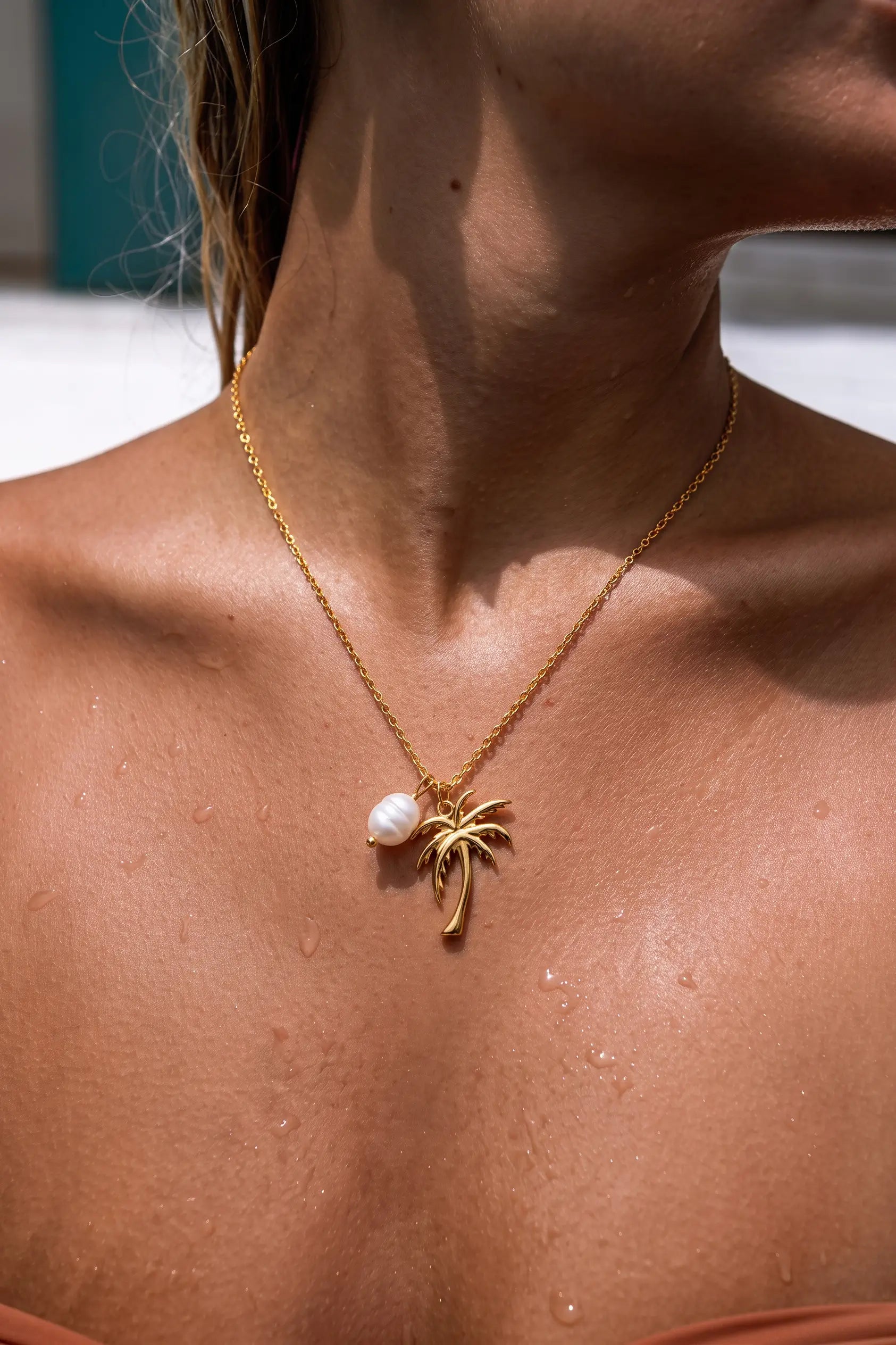 A close-up of a woman's neck and upper chest wearing a delicate gold chain necklace with a small gold palm tree pendant and a white pearl accent.