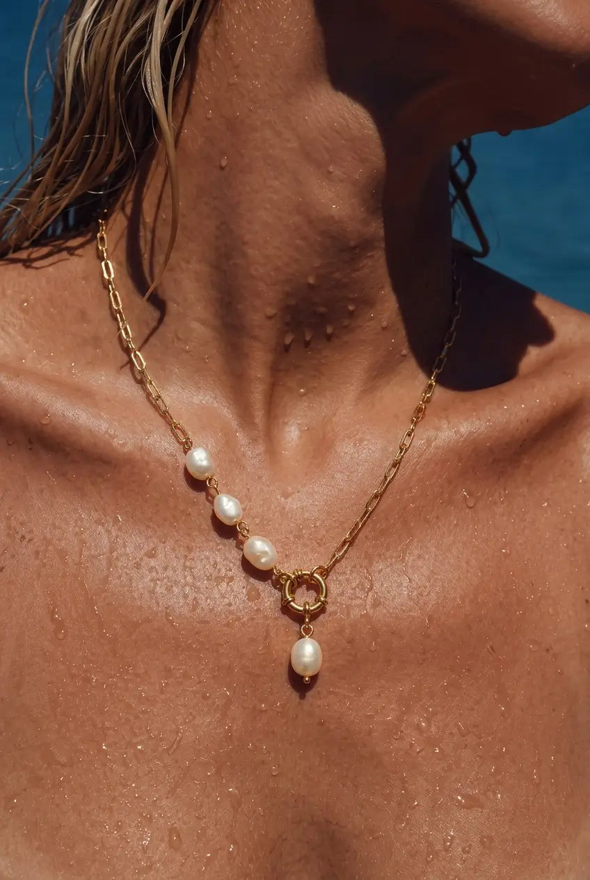 A gold chain necklace with four white freshwater pearls and a dangling pearl pendant, worn on a woman's neck against a blue water background.