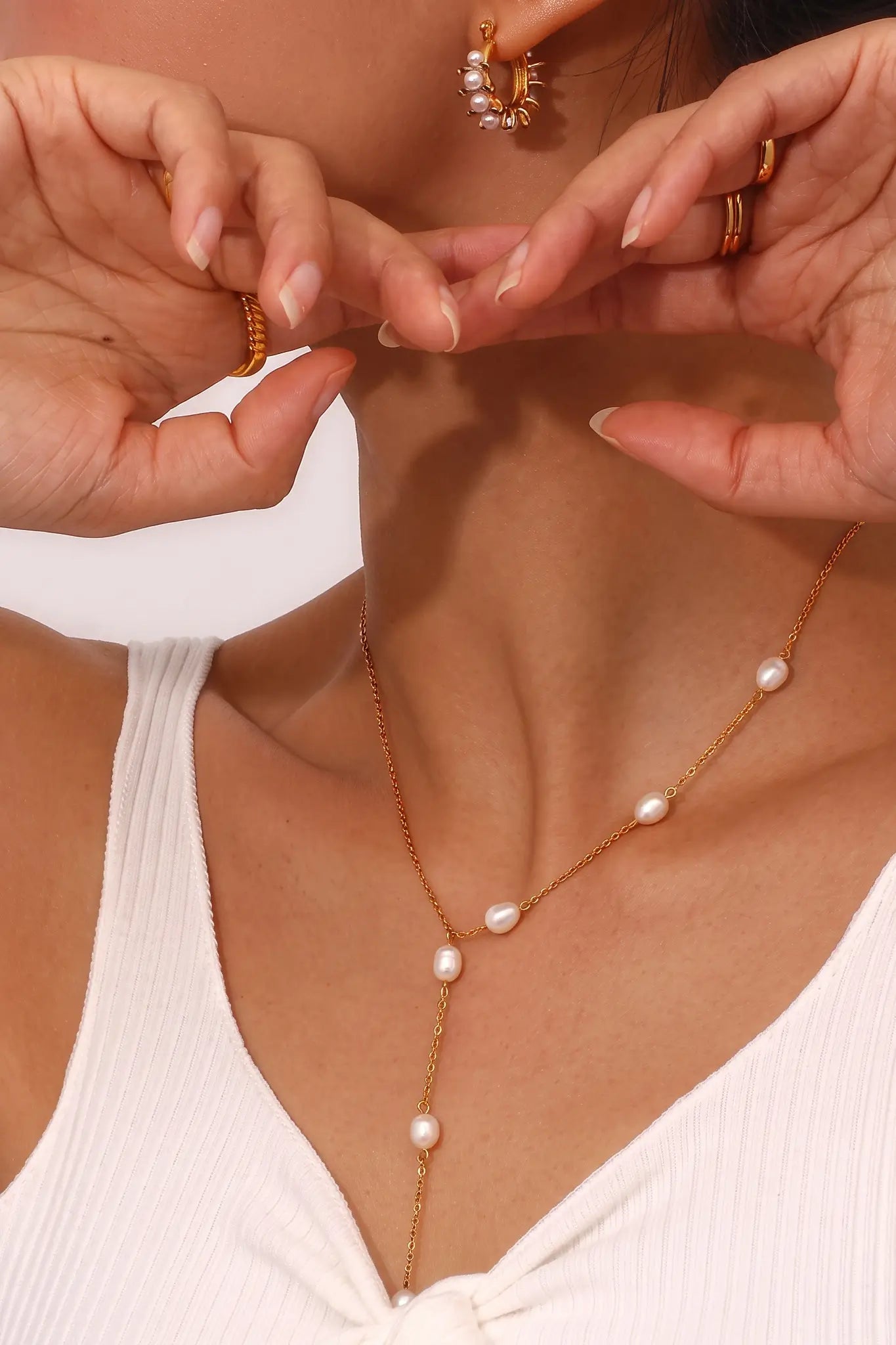 A close-up of a model wearing a delicate gold lariat necklace with white freshwater pearls, styled with matching gold hoop earrings and rings.