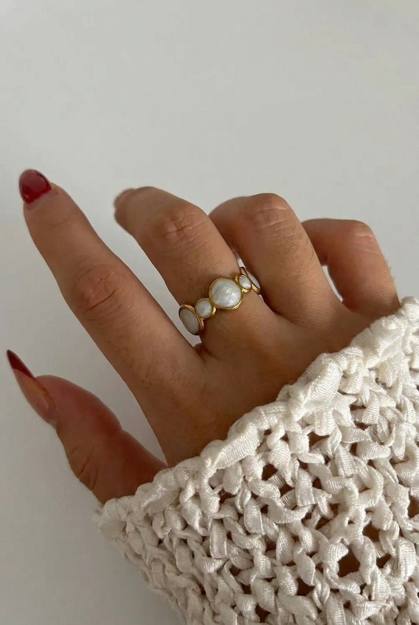 A close-up of a hand wearing a gold ring with three white pearl-like oval stones, styled with a white knit sleeve against a plain light background.