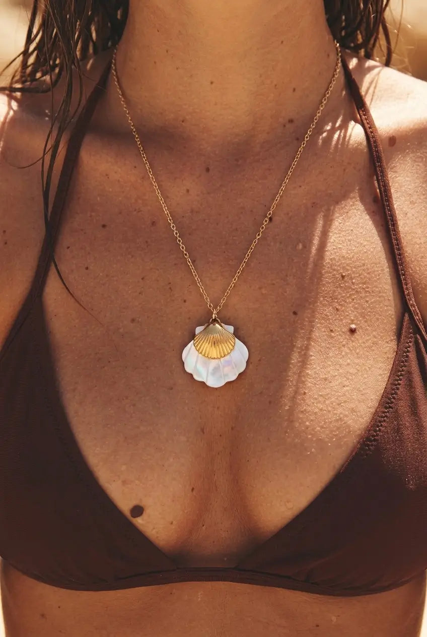 A close-up of a woman wearing a gold chain necklace with a white and gold seashell pendant, styled with a brown bikini top in warm sunlight.