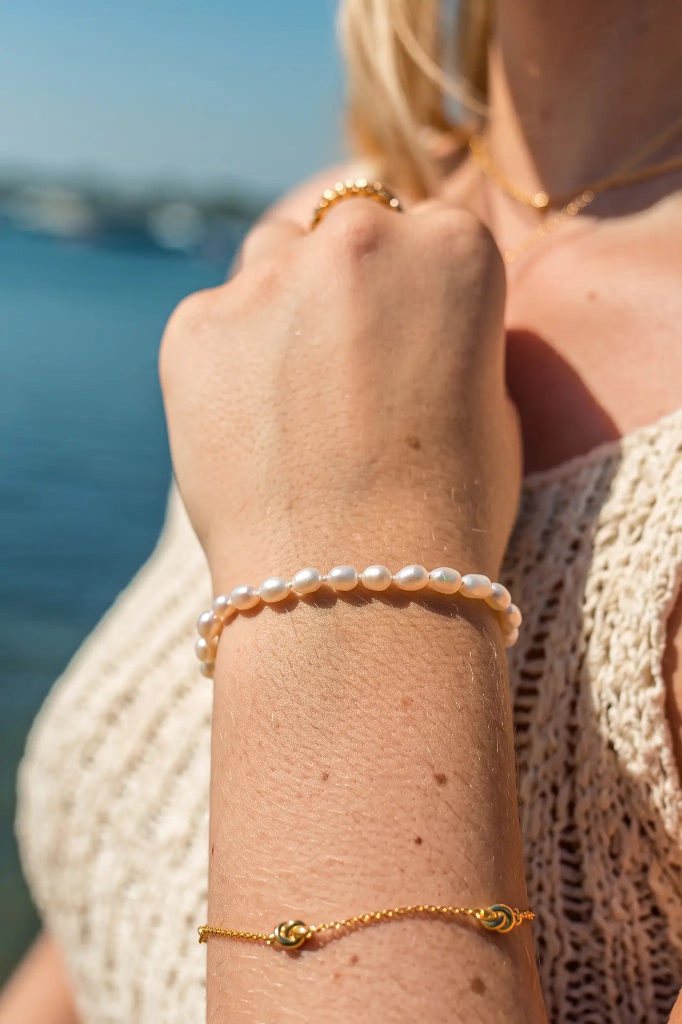 A close-up of a person's wrist wearing two delicate bracelets: a white pearl bracelet and a gold chain bracelet with a small green and gold medallion, set against a blurred background of blue water and sky.