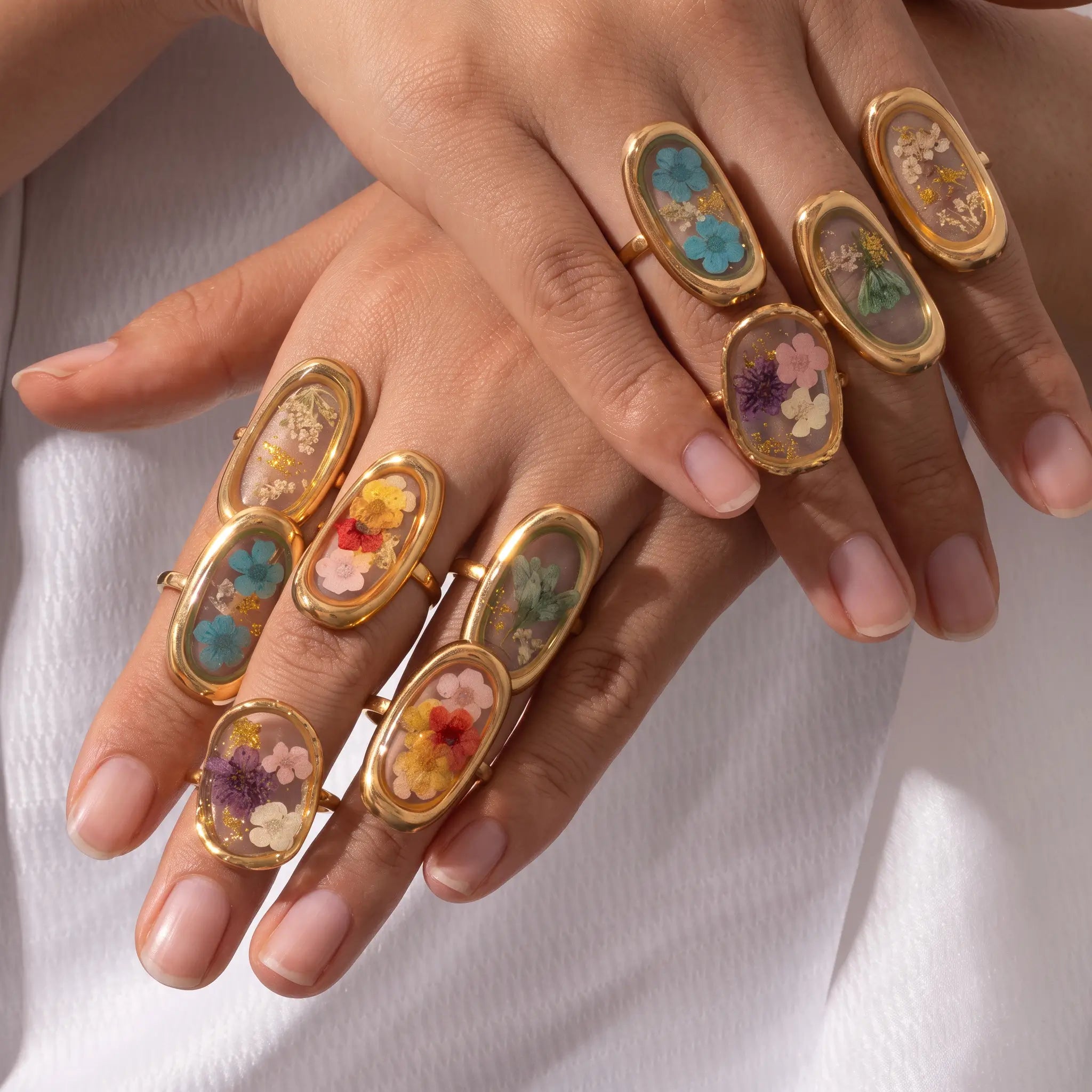 Close-up of a person's hands wearing multiple gold rings with oval inlays containing dried flowers and small specimens, set against a white fabric background.