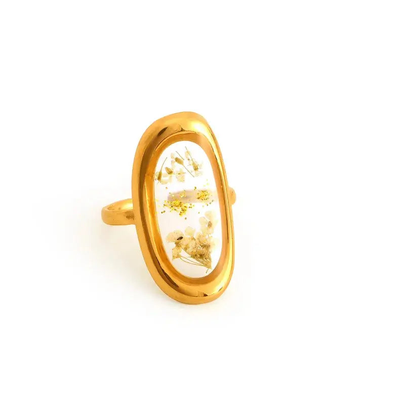 A gold oval ring featuring a clear inlay filled with dried white flowers and small gold flecks, set against a plain white background.
