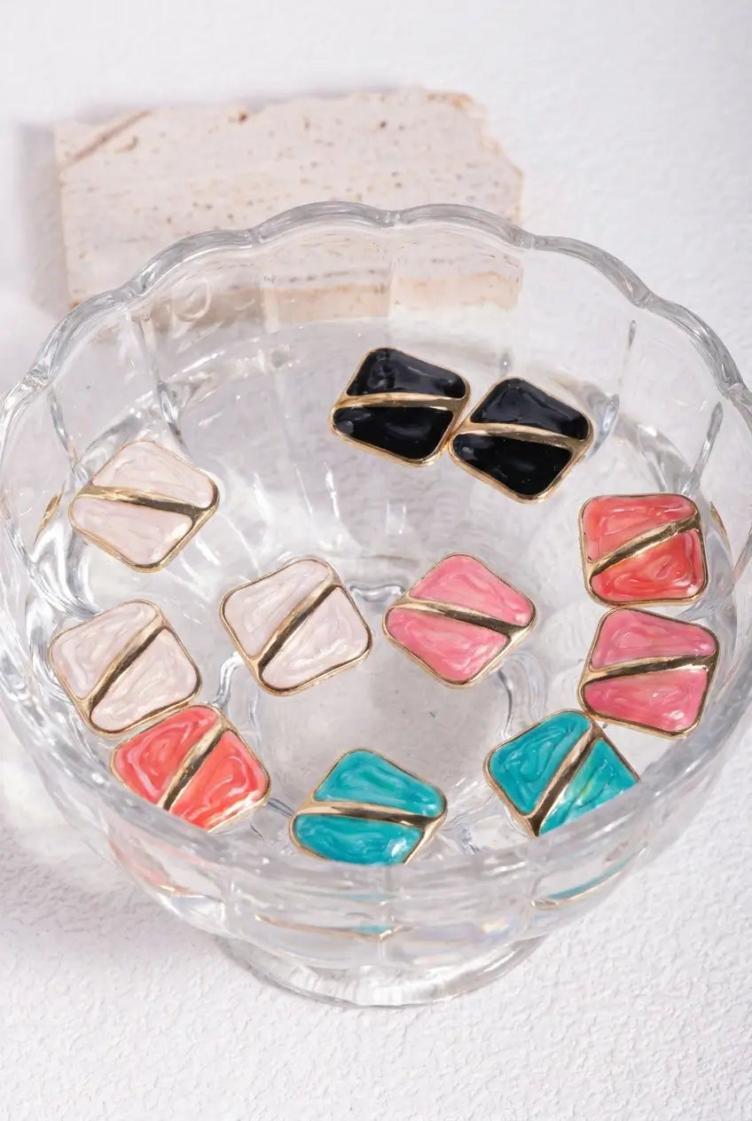 A set of colorful geometric stud earrings with gold-plated borders and multi-colored enamel centers, displayed inside a clear scalloped glass bowl on a white surface.