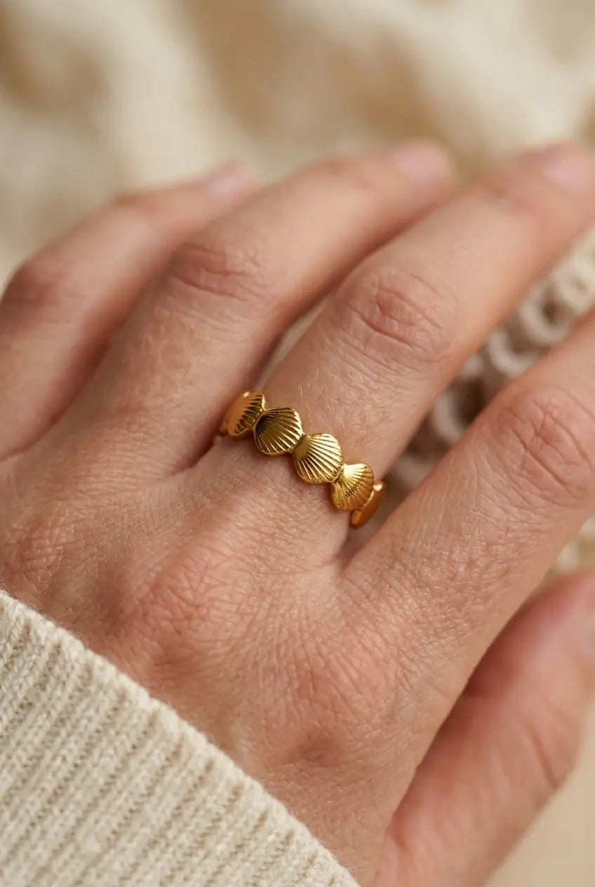 A close-up of a hand wearing a delicate gold ring with five small, textured shell-like beads, styled with a cream knit sweater against a soft, warm neutral background.