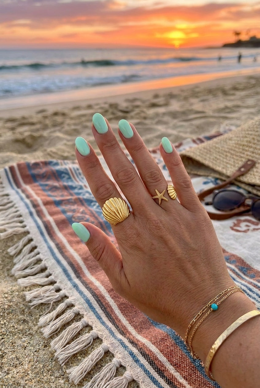 A close-up of a hand with mint green nails resting on a beach towel at sunset, wearing gold-toned beach jewelry including a seashell ring, a starfish ring, and a bangle bracelet, with sunglasses and a straw hat nearby.
