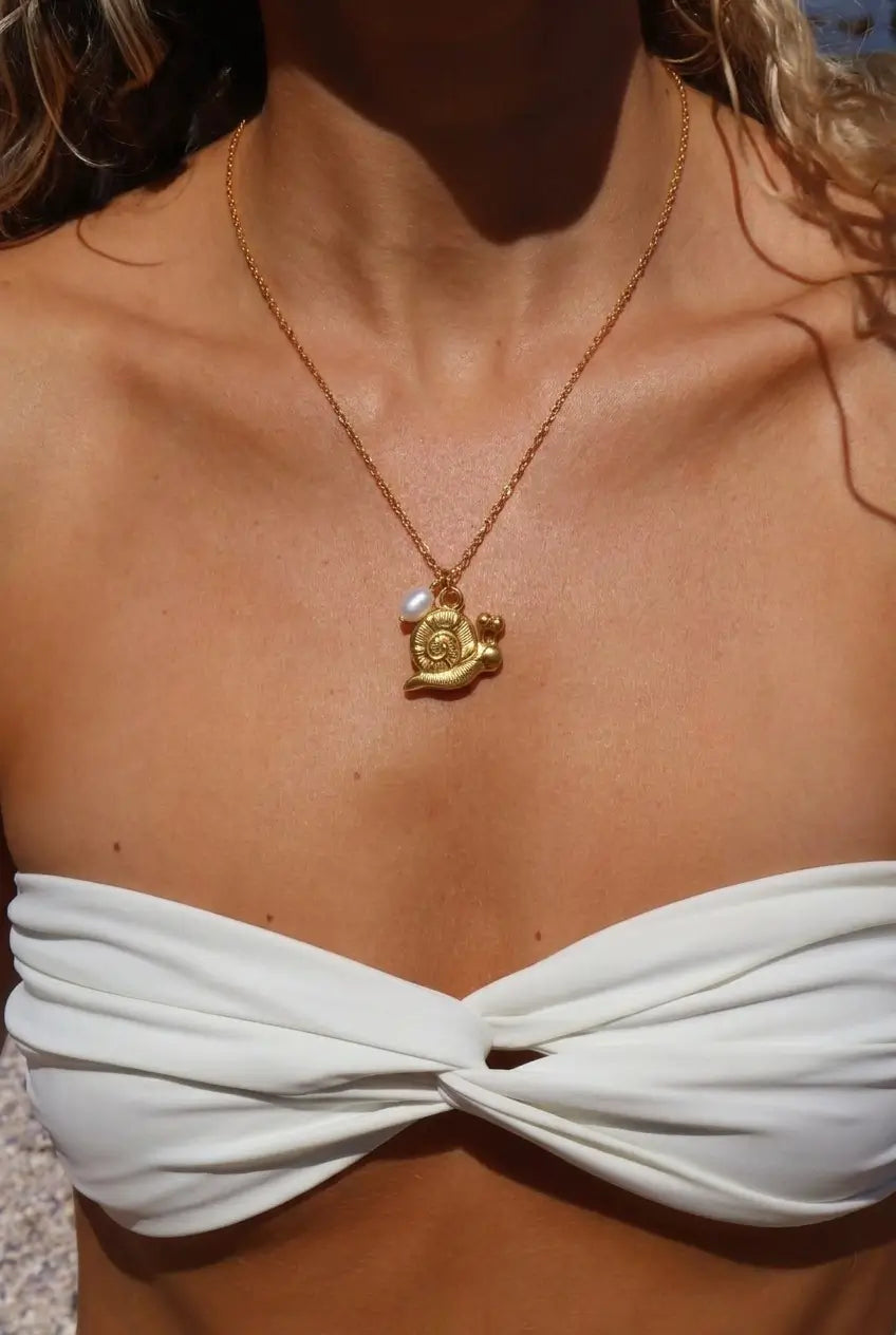 A close-up lifestyle shot of a model wearing a delicate gold chain necklace with a small gold turtle pendant and a tiny white pearl accent, styled with a white strapless bikini top.