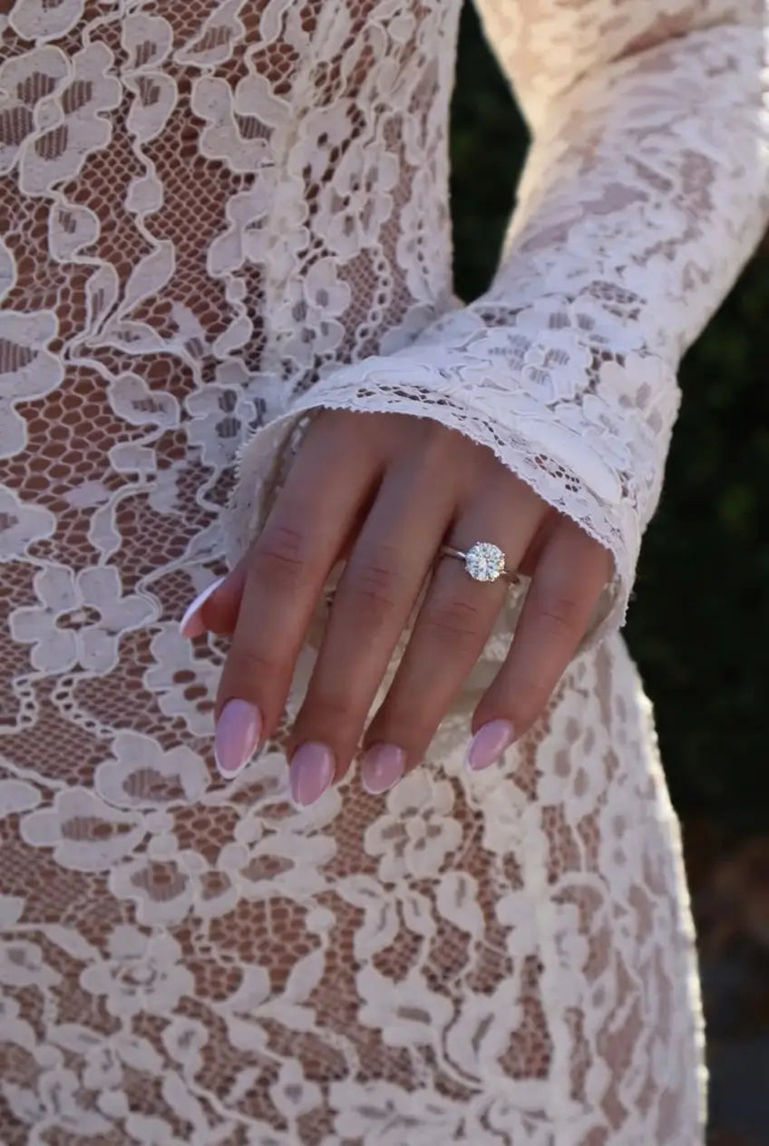 A close-up of a person's hand wearing a silver-toned ring with a round clear gemstone, shown against a white lace dress.